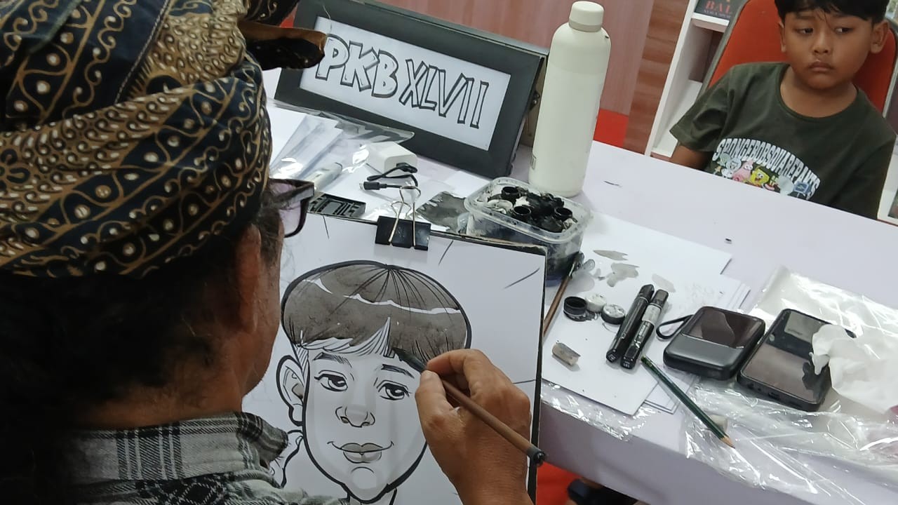 Penyempurnaan.  Wayan Tama menyempurnakan sketsa wajah kartun seorang anak. Ia menyelesaikannya secara hati-hati dengan hati dengan memoleskan kuasnya tipis-tipis yang telah dicelupkan tinta warna hitam.